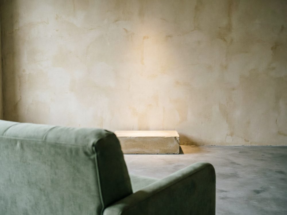 Sofá verde com parede bege texturizada e chão cinza num ambiente minimalista