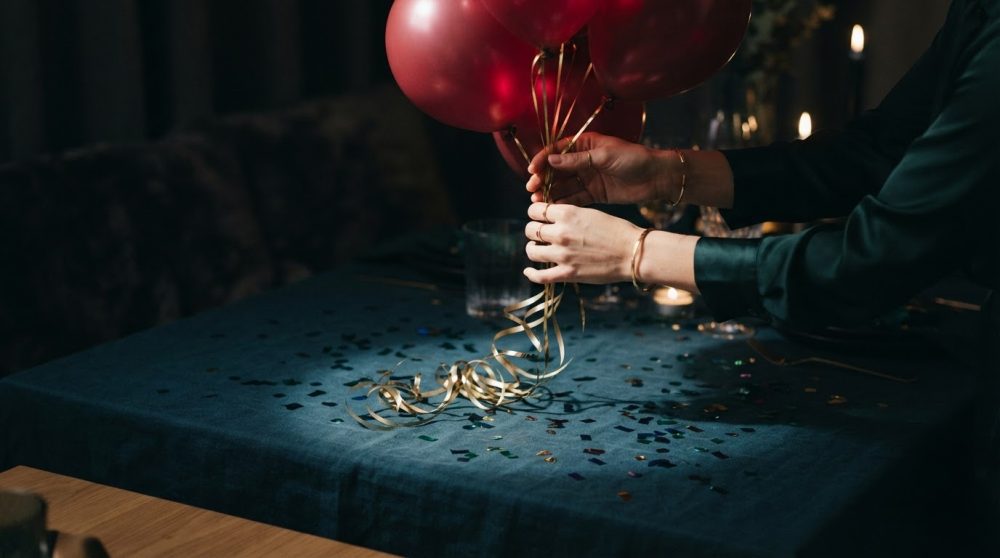 Mãos segurando balões vermelhos com fitas douradas sobre mesa azul com confetis