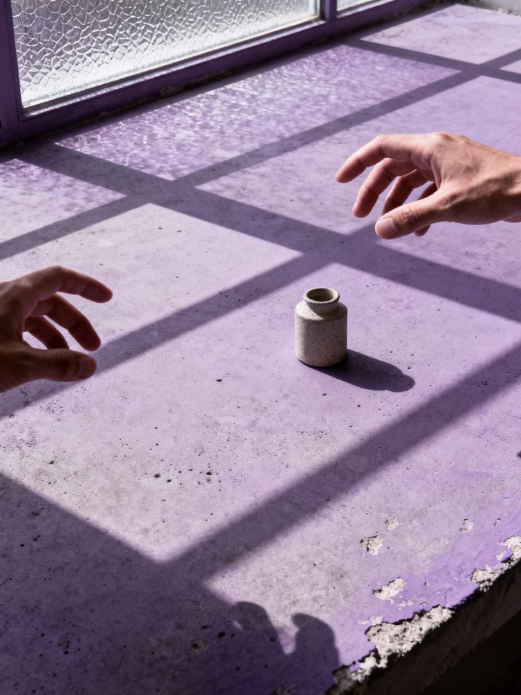 Superfície roxa desgastada com pequeno frasco branco e sombras de mãos junto a janela