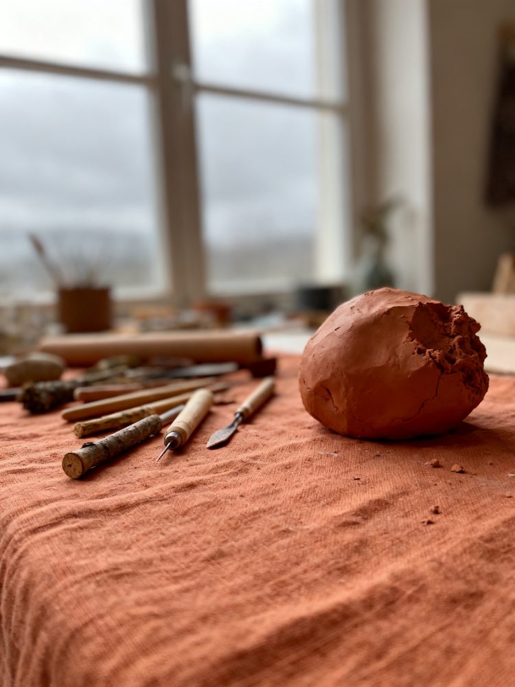 Mesa com bola de barro e ferramentas de modelagem artesanal junto a uma janela com luz natural