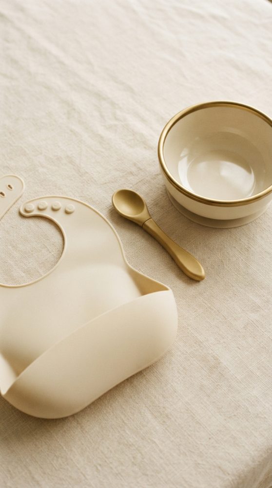 Bibe de silicone beige, colher dourada e taça creme sobre tecido claro