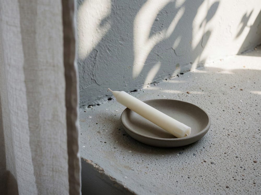 Vela branca deite num prato de cerâmica sobre superfície texturada com sombras de plantas
