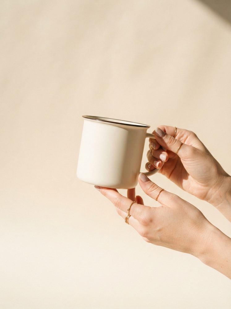 Mãos segurando caneca metálica branca com borda cromada sobre fundo bege