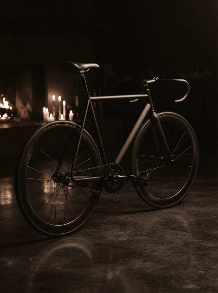 Bicicleta fixa preta com rodas largas perto de lareira e velas acesas num ambiente escuro