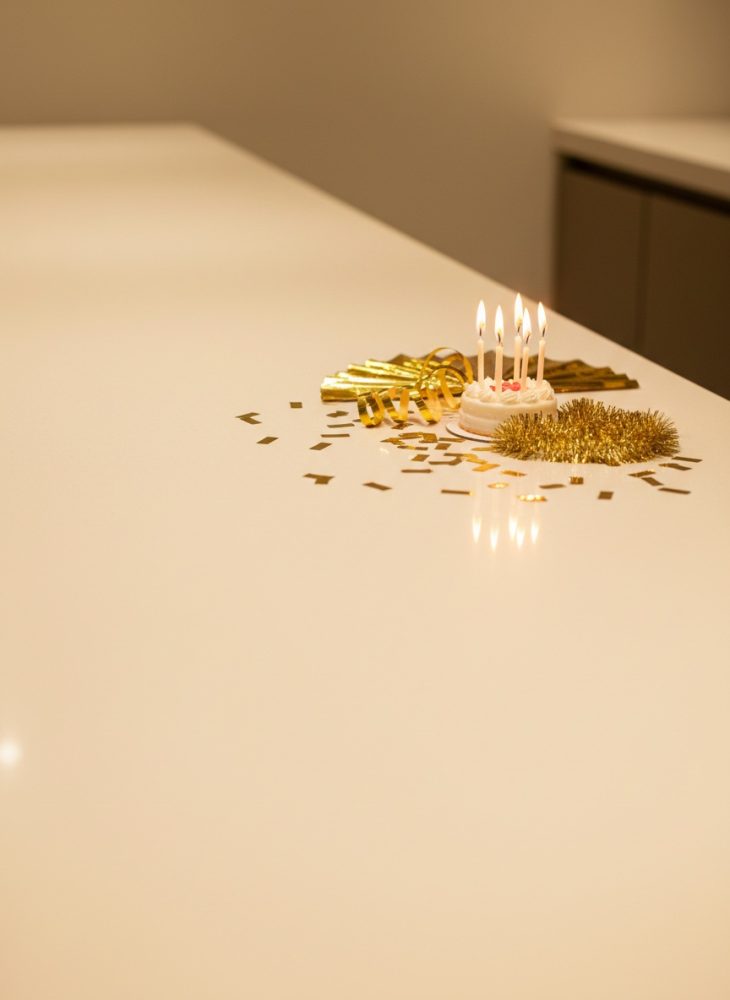 Mini bolo branco com cinco velas acesas e decorações douradas sobre mesa branca