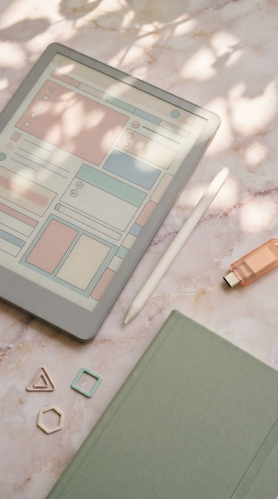 Tablete digital, caneta, pen USB, caderno verde e clips geométricos sobre mármore com sombras suaves