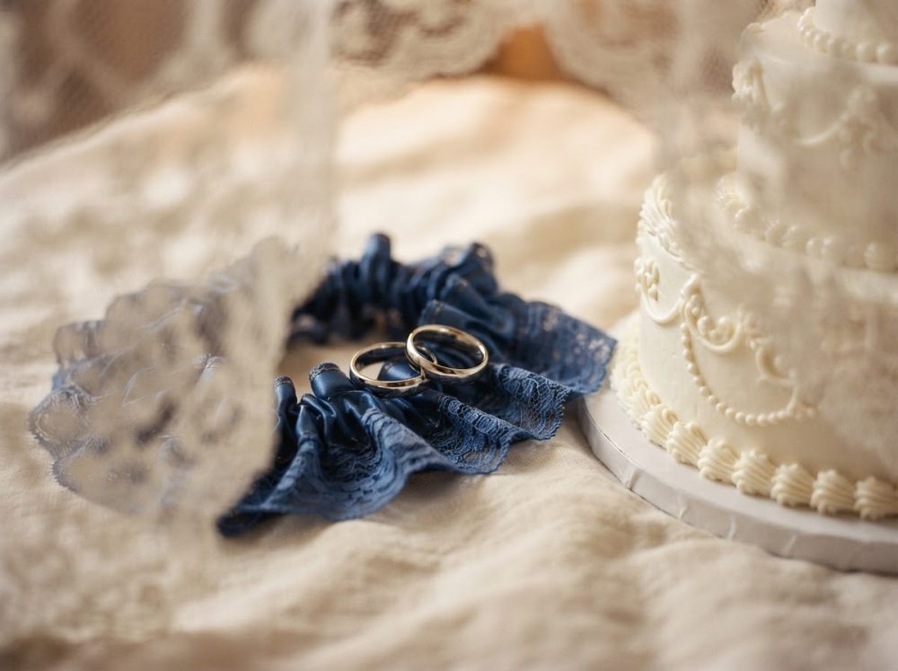Alianças douradas sobre liguete azul e bolo de casamento branco detalhado