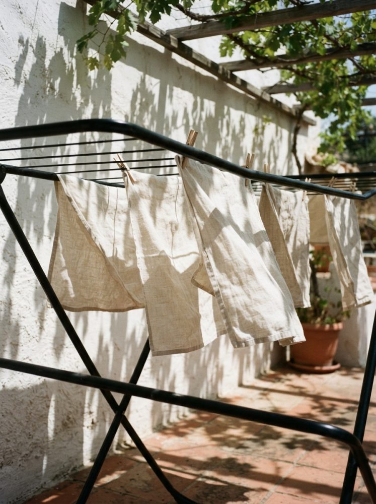 Varal de roupa com panos claros a secar sob sombra de planta numa varanda ensolarada
