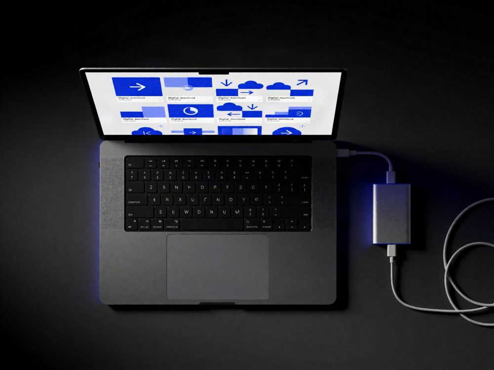 Computador portátil cinzento com ecrã aberto e drive externa ligada por cabo numa superfície escura