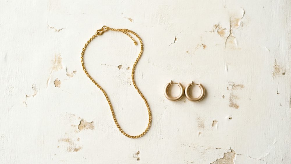 Colar dourado com corrente fina e brincos de argola dourados sobre fundo branco texturado