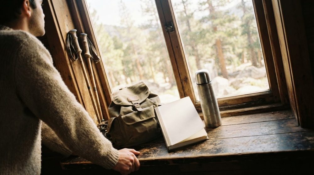 Homem de suéter bege observa mochila, bastões, cantil e livro aberto numa janela rústica com vista para floresta