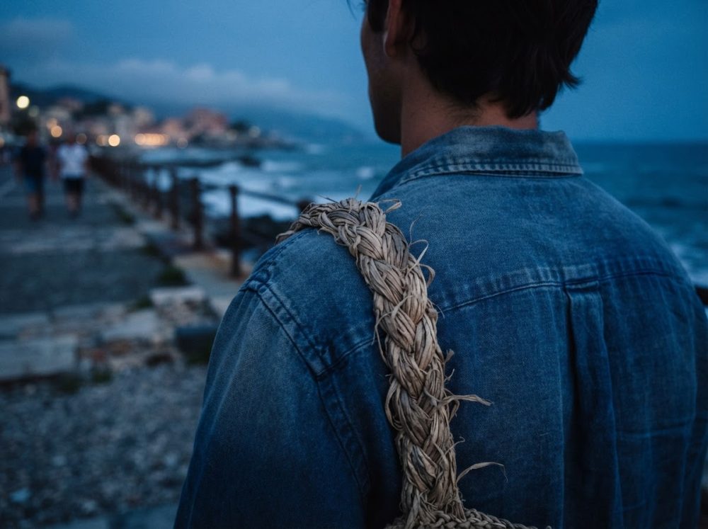 Pessoa de costas com jaqueta de ganga azul e saco trançado de corda perto do mar ao entardecer