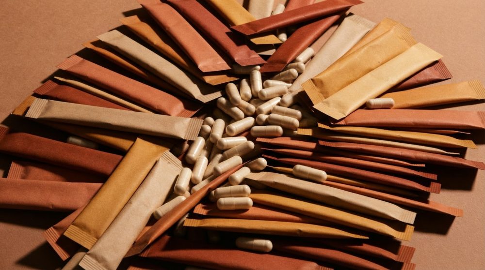 Pacotes individuais de suplemento em papel kraft e cápsulas brancas sobre fundo castanho