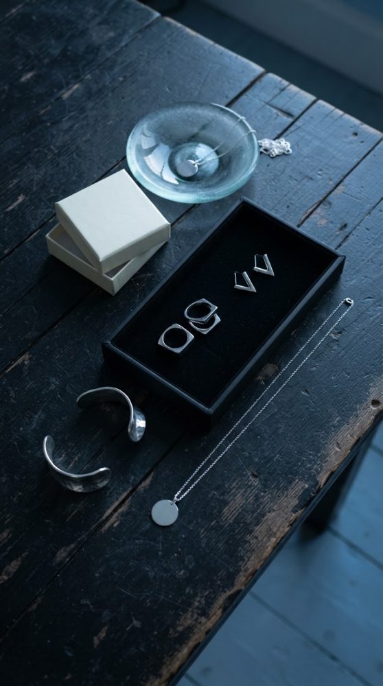 Joias em prata minimalistas como pulseira, colar, anéis e brincos sobre mesa de madeira escura