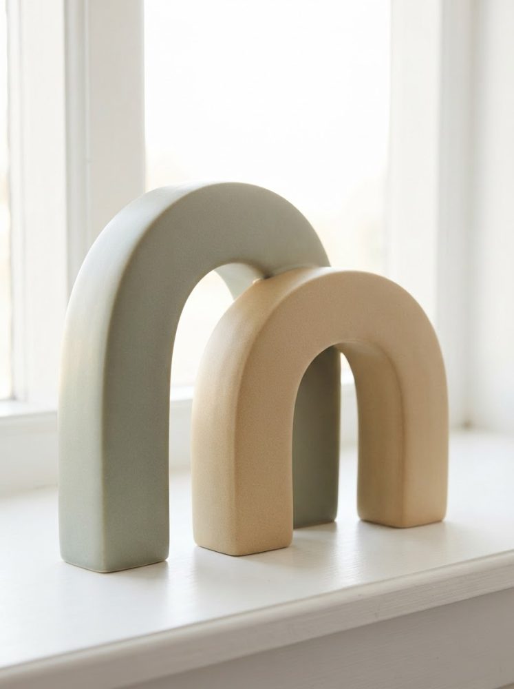 Duas esculturas decorativas em forma de arco, uma verde e outra bege, sobre uma superfície branca junto a uma janela.