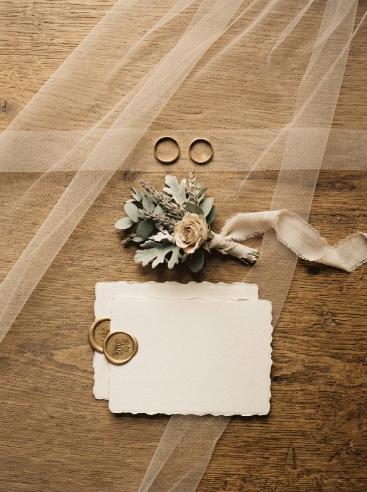 Convites de casamento com selo de cera, arranjo floral, anéis dourados e tule sobre madeira