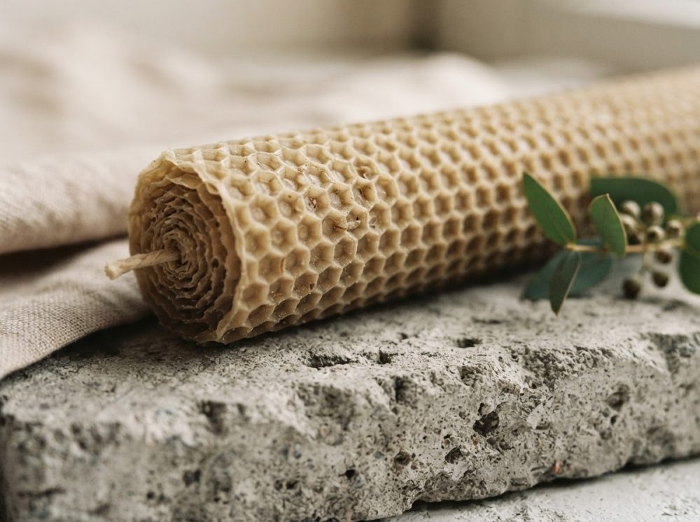 Vela artesanal de cera de abelha com padrão hexagonal sobre pedra rugosa e folhas verdes