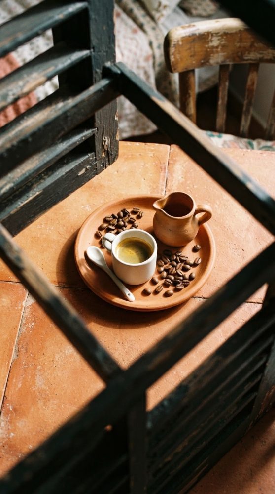 Xícara de café expresso num prato de barro com grãos de café e jarra pequena sobre mesa de azulejo