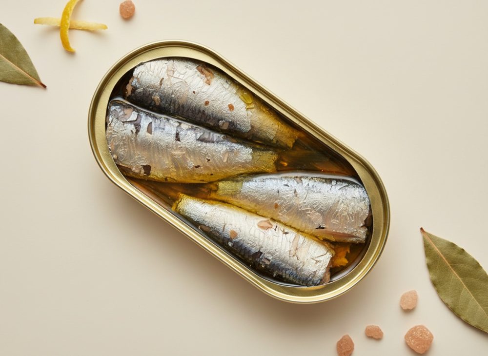 Lata aberta de sardinhas em azeite com folhas de louro e cristais de sal rosa num fundo claro
