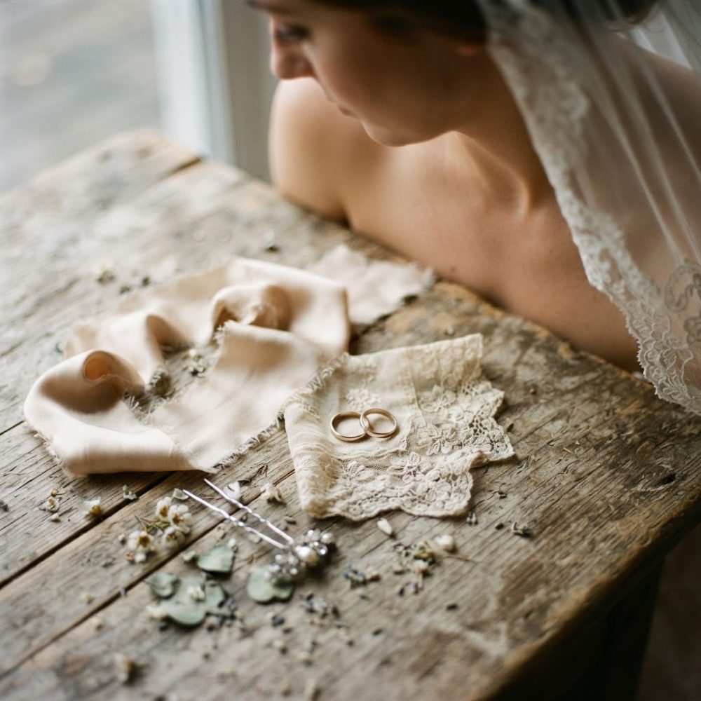Alianças de casamento sobre renda e tecido com acessórios numa mesa rústica vista de cima