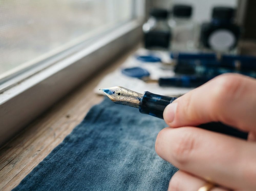 Mão segurando caneta tinteiro azul com frascos de tinta ao fundo em superfície de madeira