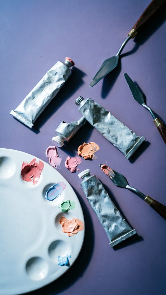 Tubos de tinta acrílica, paleta com tintas coloridas e espátulas sobre fundo roxo
