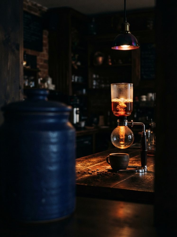 Café a ser preparado num sifão sobre bancada de madeira, ambiente rústico e luz quente