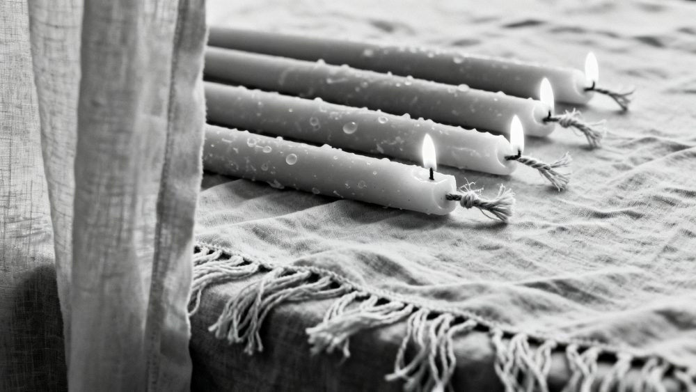 Quatro velas brancas finas acesas alinhadas sobre tecido com franja em ambiente calmo