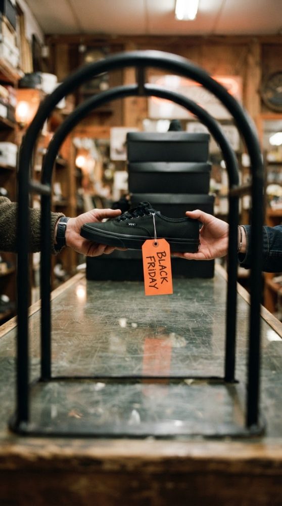 Mãos trocando ténis preto com etiqueta Black Friday numa loja com caixas ao fundo
