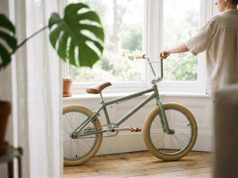 Bicicleta infantil verde pastel junto a janela iluminada, com piso de madeira e plantas ao redor