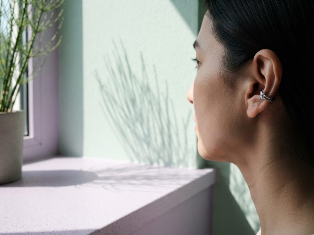 Mulher de perfil com brincos de prata junto a janela com plantas e sombra suave
