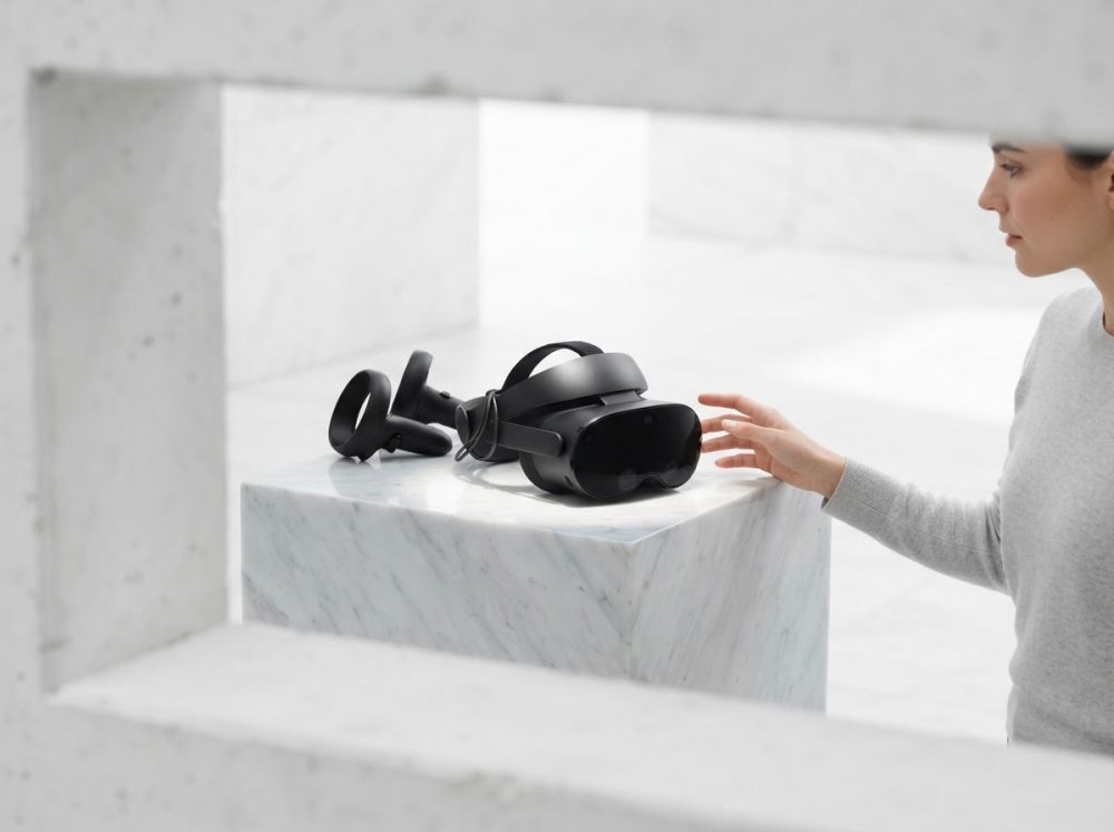 Headset de realidade virtual preto com dois controladores sobre suporte branco, e mulher a tocar no headset.
