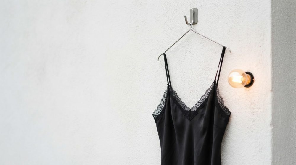 Camisola preta com renda pendurada num cabide metálico contra parede branca com lâmpada incandescente