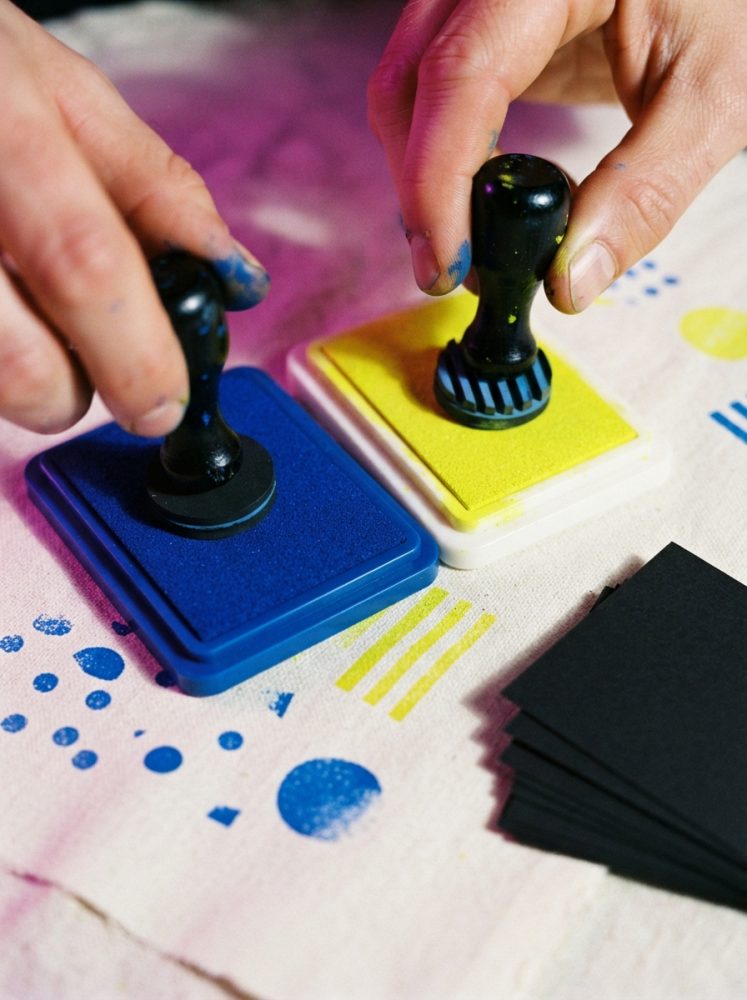 Mãos seguram carimbos em almofadas de tinta azul e amarela sobre papel branco com marcas coloridas