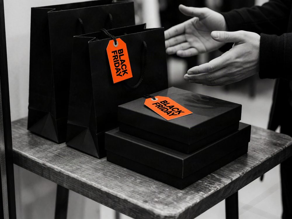 Sacos e caixas pretas com etiquetas laranja Black Friday sobre mesa de madeira e mãos ao lado