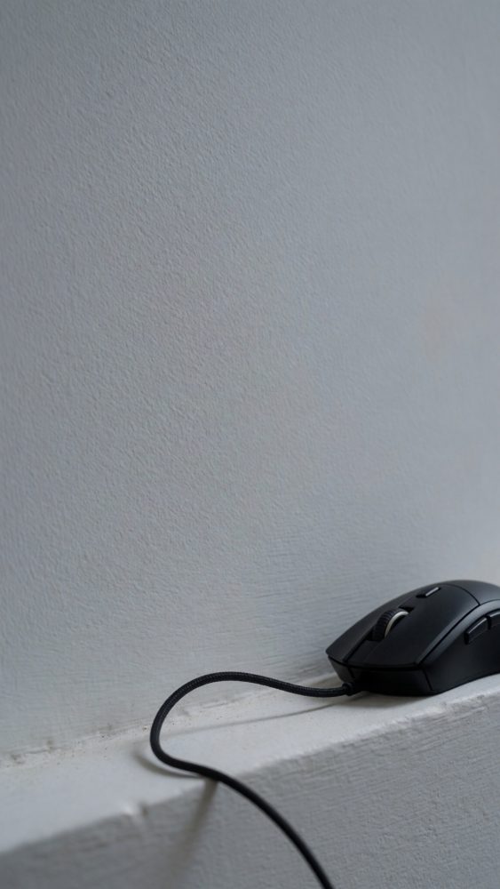 Rato negro gaming com cabo sobre superfície branca e parede cinzenta
