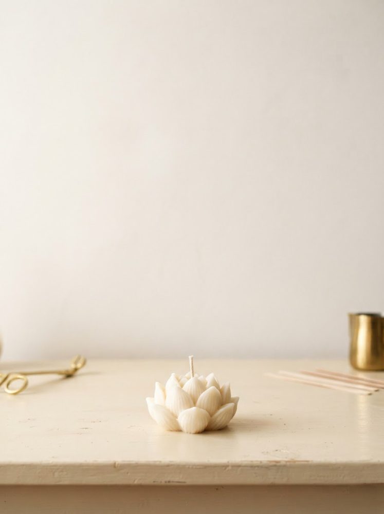 Vela branca em forma de flor de lótus sobre uma mesa clara com objetos decorativos dourados ao fundo