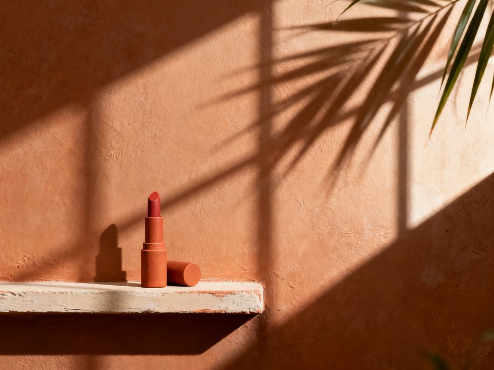 Batom vermelho sobre prateleira branca com sombra de folhas e luz natural em fundo laranja texturado