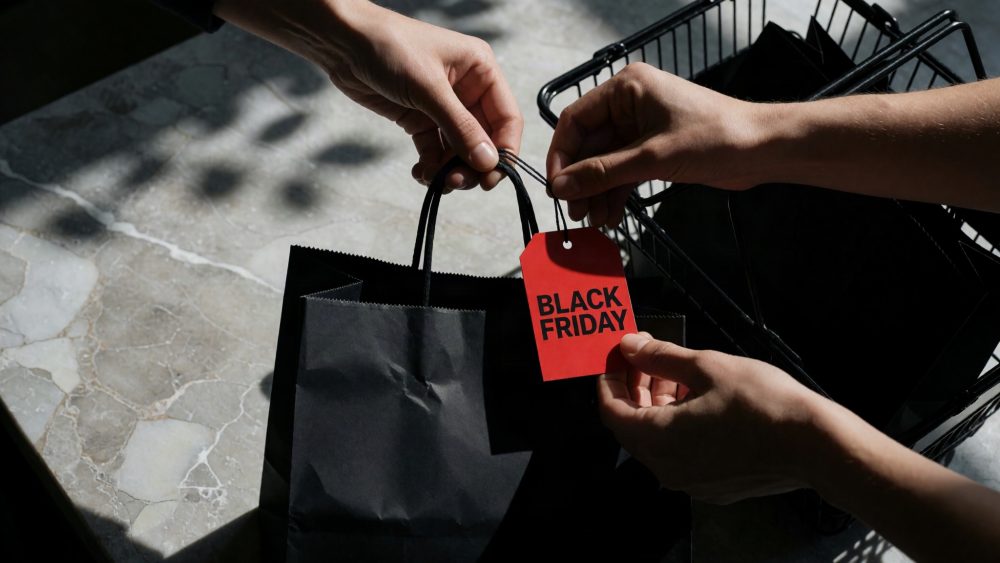 Saco de compras preto com etiqueta vermelha de Black Friday e mãos segurando-o sobre chão de pedra