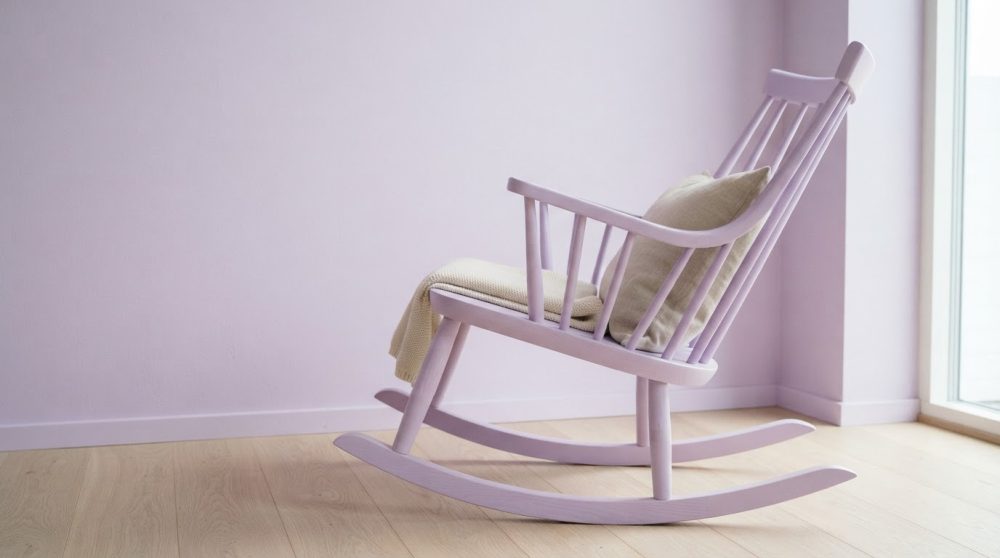 Cadeira de baloiço lilás com almofada e manta num quarto com parede lilás e chão em madeira clara
