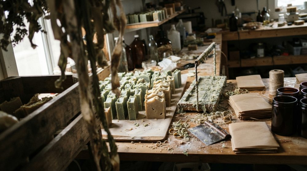 Mesa rústica com sabonetes artesanais, ferramentas de corte e embalagens em ambiente de produção