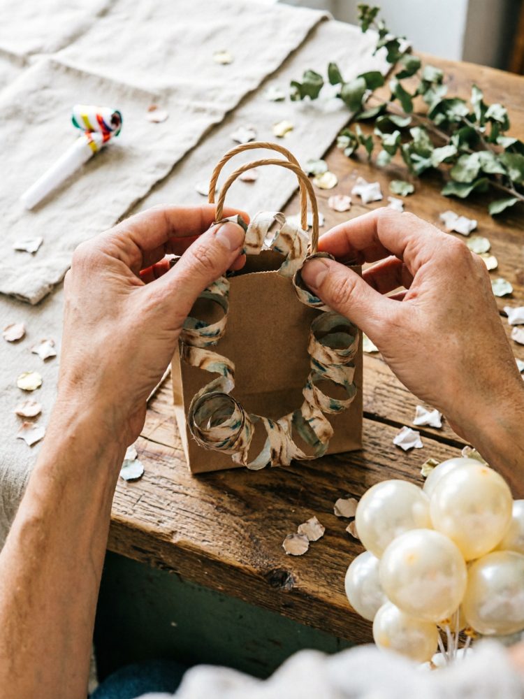 Mãos a decorar saco de papel com serpentinas, confetes e balões brancos sobre mesa de madeira