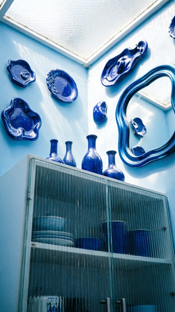Parede azul com decoração em cerâmica azul e espelho ondulado acima de armário de vidro