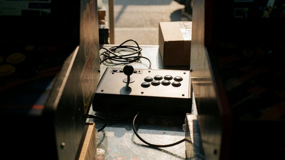 Joystick arcade preto com oito botões iluminado pela luz natural entre máquinas arcade