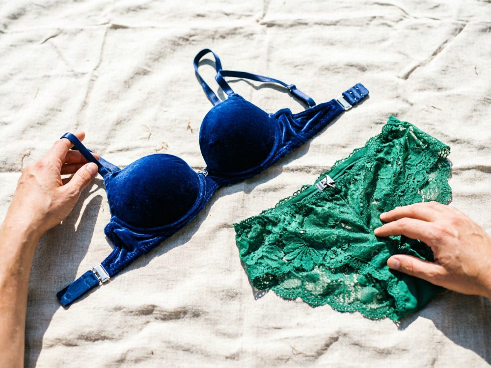Conjunto lingerie com sutiã azul veludo e cueca verde renda sobre tecido claro com mãos a tocar