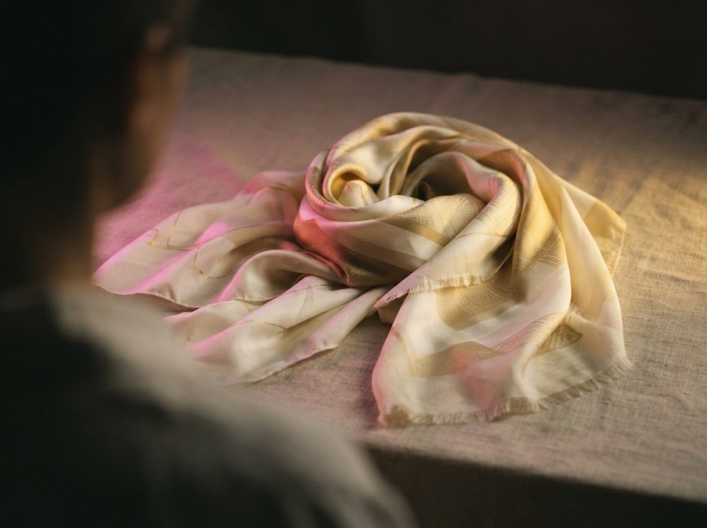 Lenço de seda bege e dourado enrolado sobre tecido de textura rústica com luz suave rosa e amarela