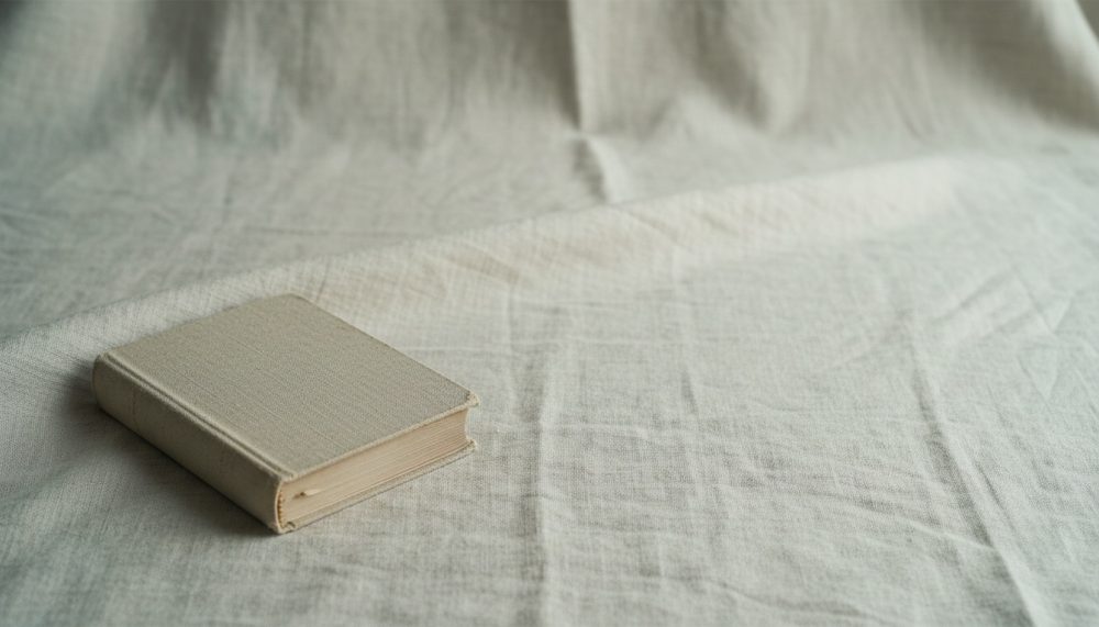 Livro antigo com capa bege repousa sobre pano texturado da mesma cor