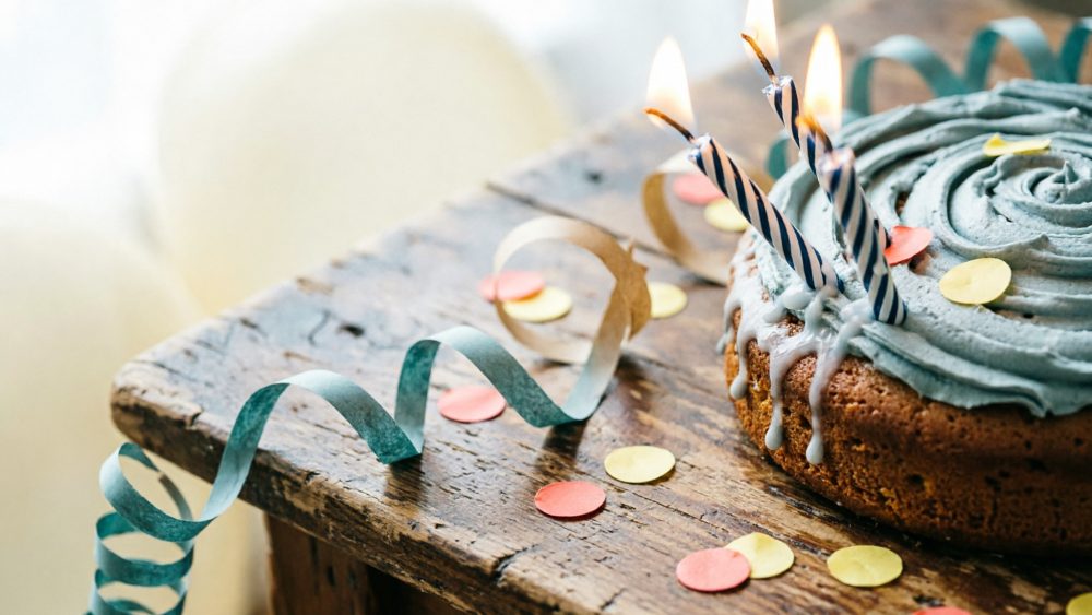 Bolo azul com três velas acesas sobre mesa de madeira com confetes e serpentinas