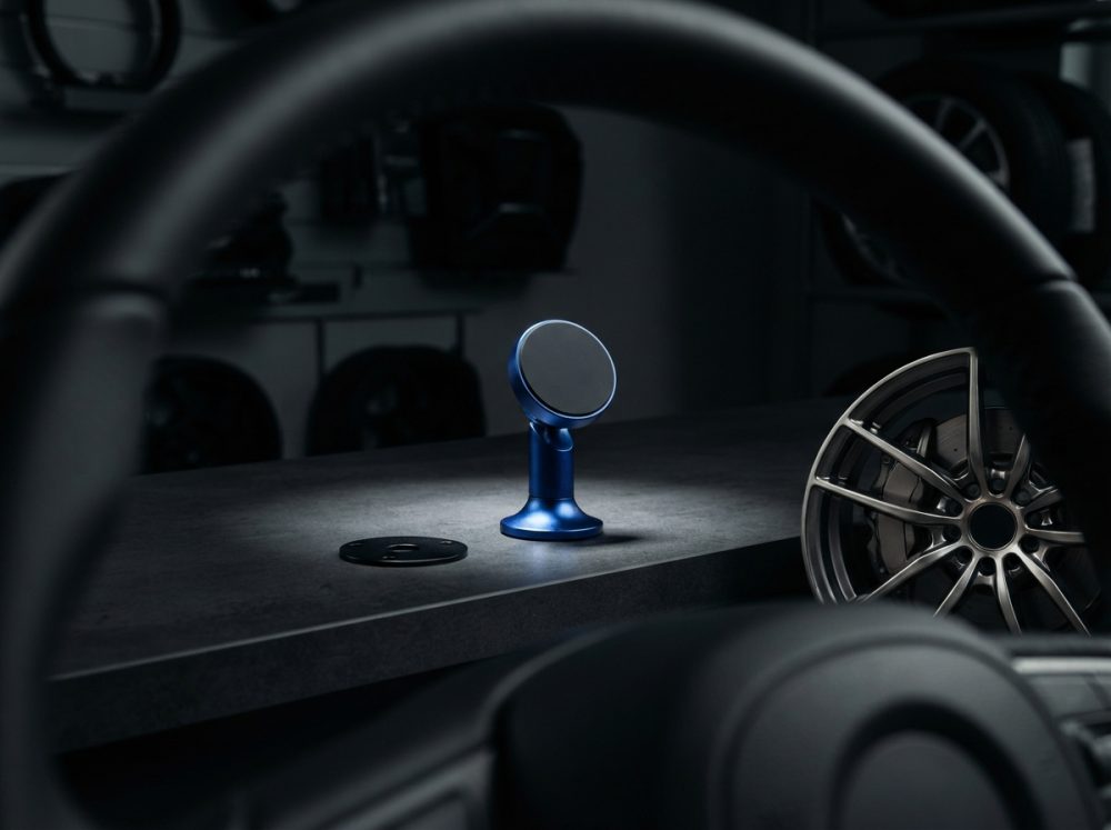 Suporte magnético azul para telemóvel numa bancada cinzenta com volante e jante automóvel ao fundo