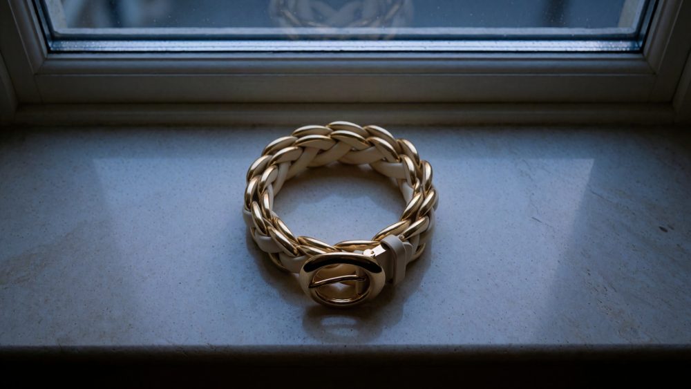Cinto trançado dourado e bege colocado numa superfície clara junto a uma janela
